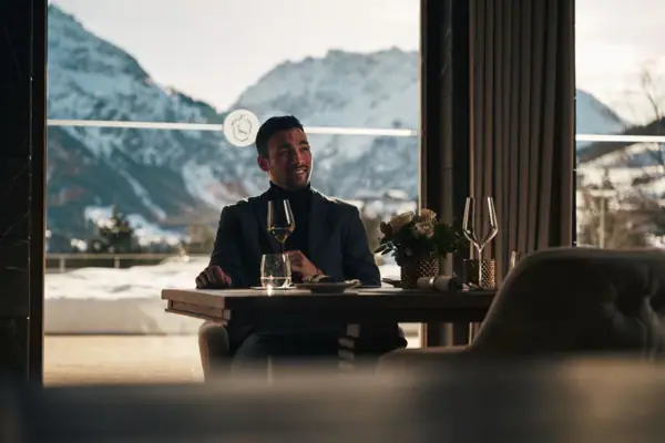 Ein Mann sitzt an einem Tisch mit Weingläsern und Bergen im Hintergrund.
