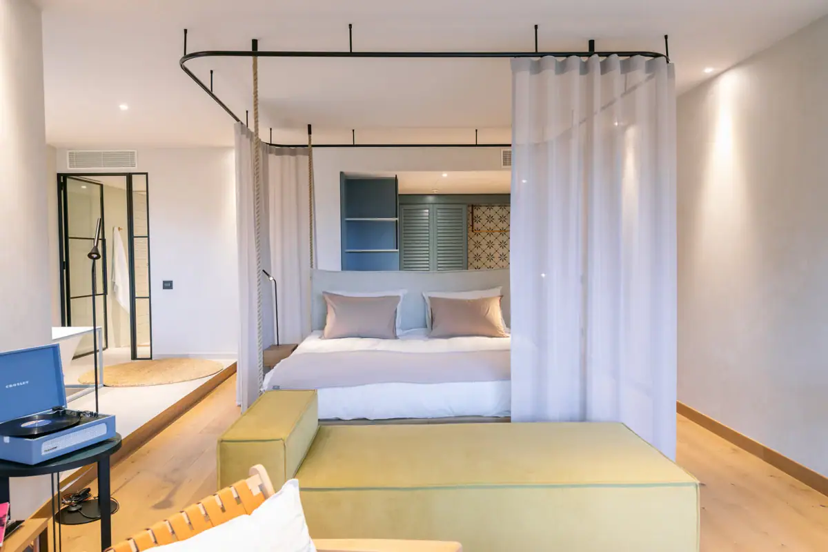 Ein Bild von einer Juniorsuite mit einem Bett. Um das Bett ist ein Vorhang, den man zuzuiehen kann. Vor dem Bett steht eine Couch. Im Hintergrund sieht man eine freistehende Badewanne. 