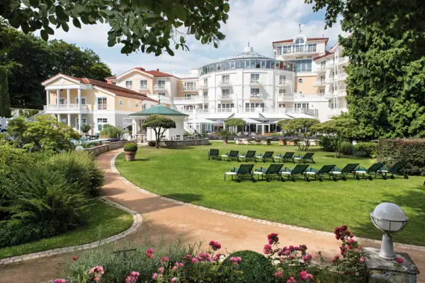 Liegestühle im Garten mit Rasen und einem Hotel sowie Bäumen im Hintergrund.
