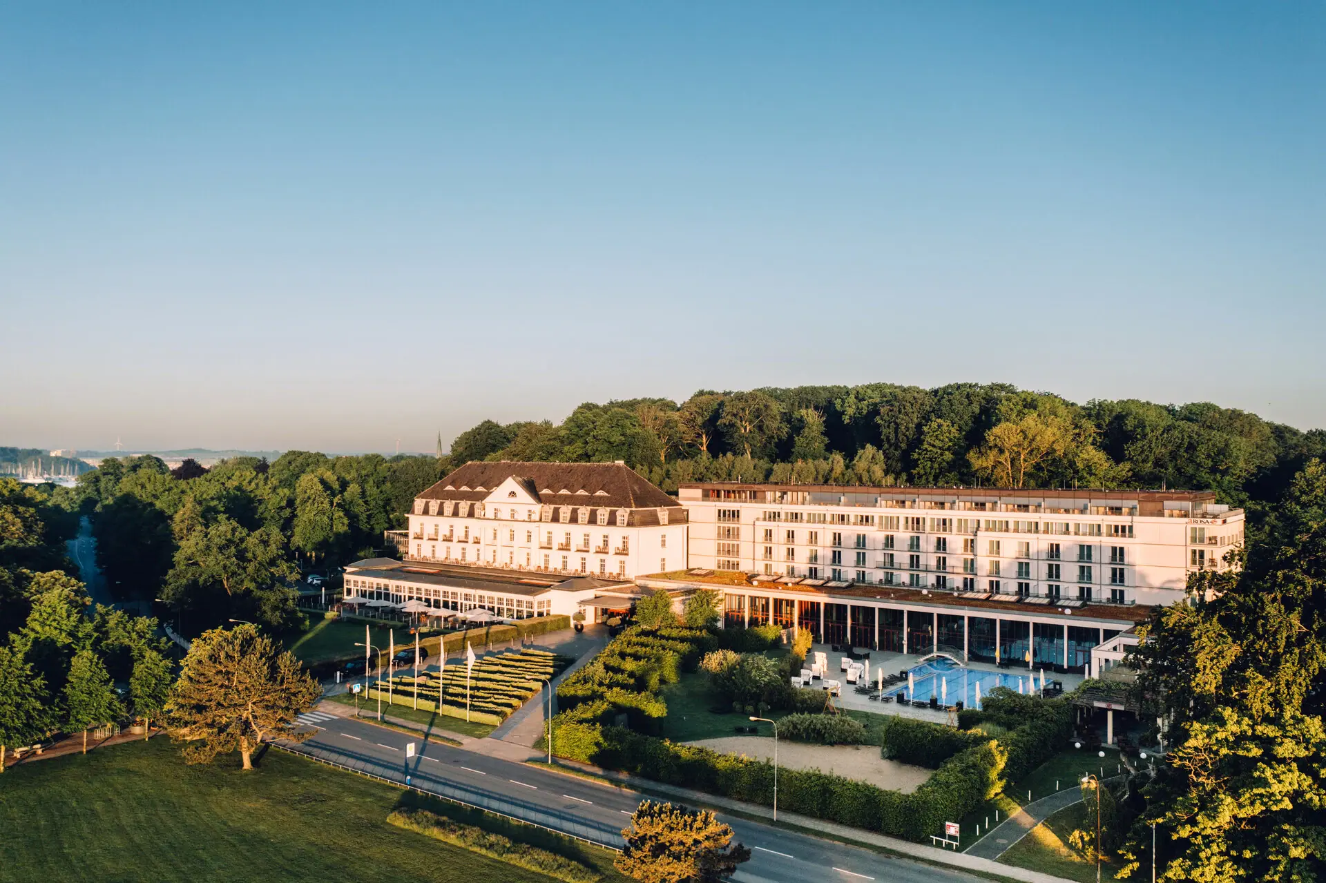 Großes weißes Gebäude mit Pool im Vordergrund und Bäumen im Hintergrund.