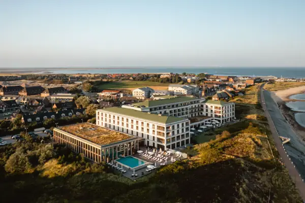 Eine Luftansicht des A-ROSA Sylt. Das helle Gebäude liegt in der grünen Landschaft und ist nur durch einen Weg vom Meer in der rechten Seite des Bildes getrennt. 