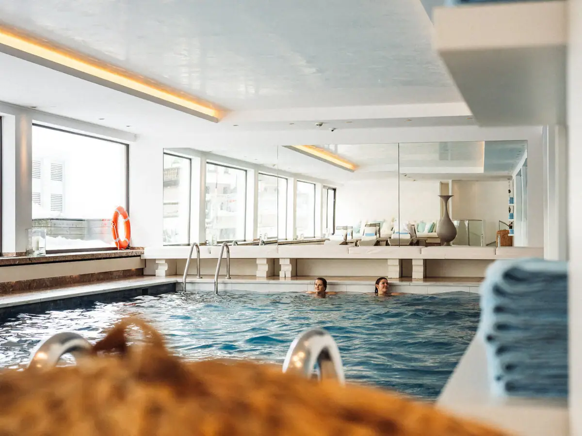 Personen im Pool vom Thunher’s Alpenhof mit großem Fenster. auf der linken Seite