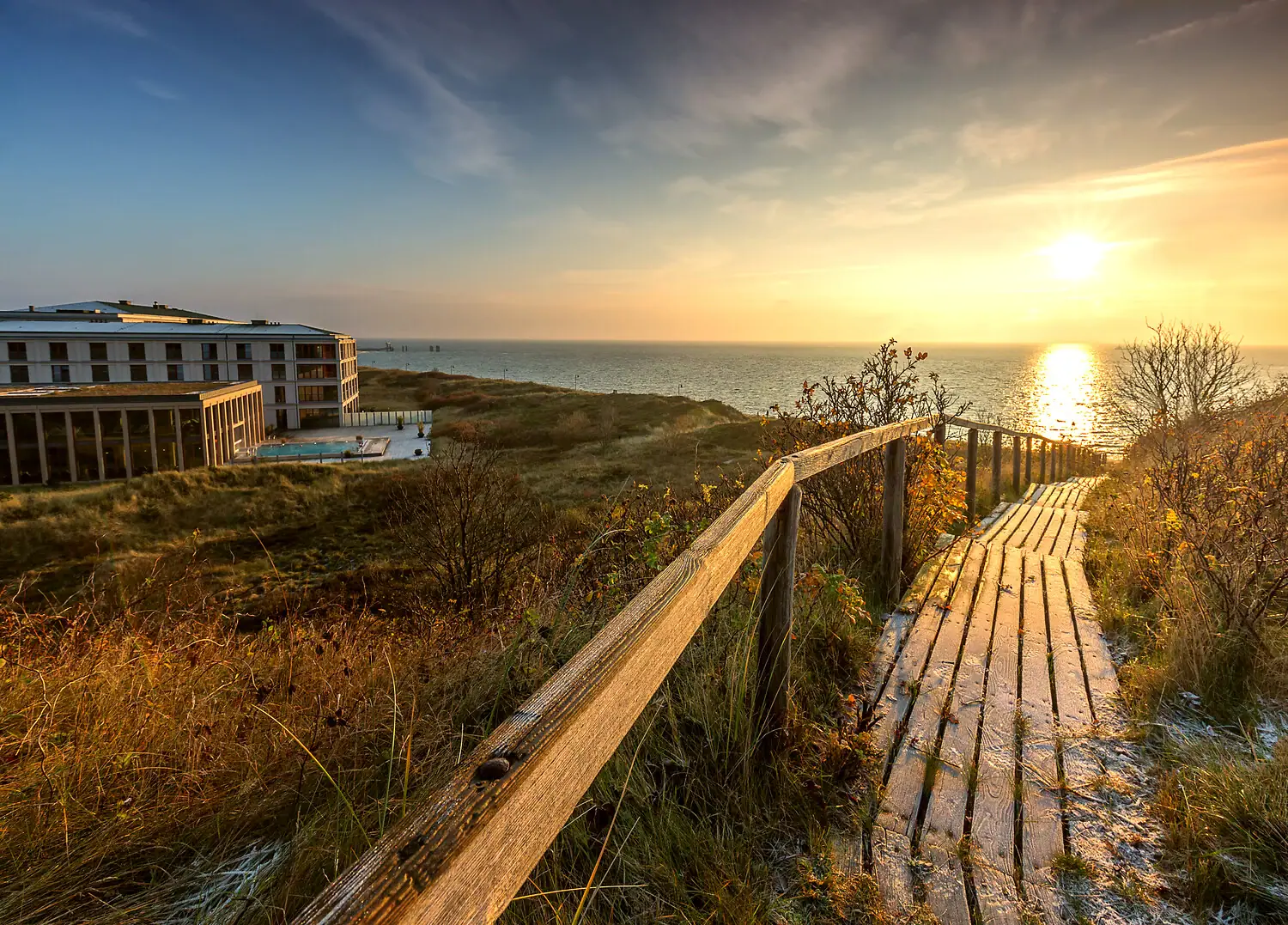 A-ROSA Sylt Holzsteg führt zum Strand mit Gebäude und Gewässer im Hintergrund.