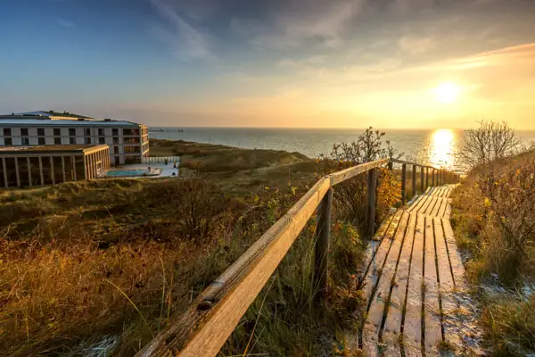 A-ROSA Sylt Holzsteg führt zum Strand mit Gebäude und Gewässer im Hintergrund.