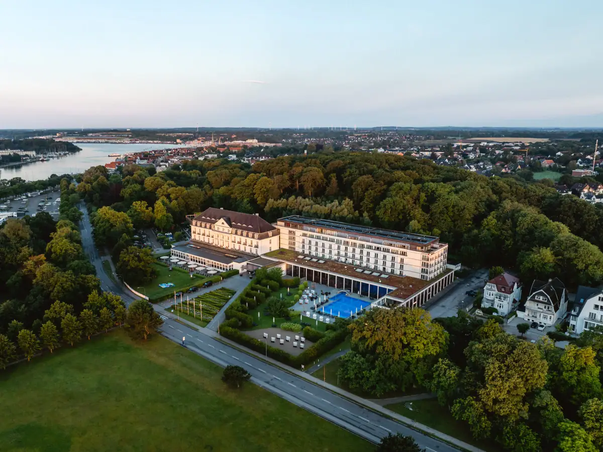 Das A-ROSA Travemünde mit Pool, umgeben von Bäumen und im Hintergrund ist die Mündung der Trave zu sehen..