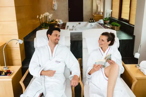 Ein Mann und eine Frau in weißen Bademänteln liegen in einem gemütlichen SPA Bereich und entspannen sich, die Frau hält ein Buch in den Händen.