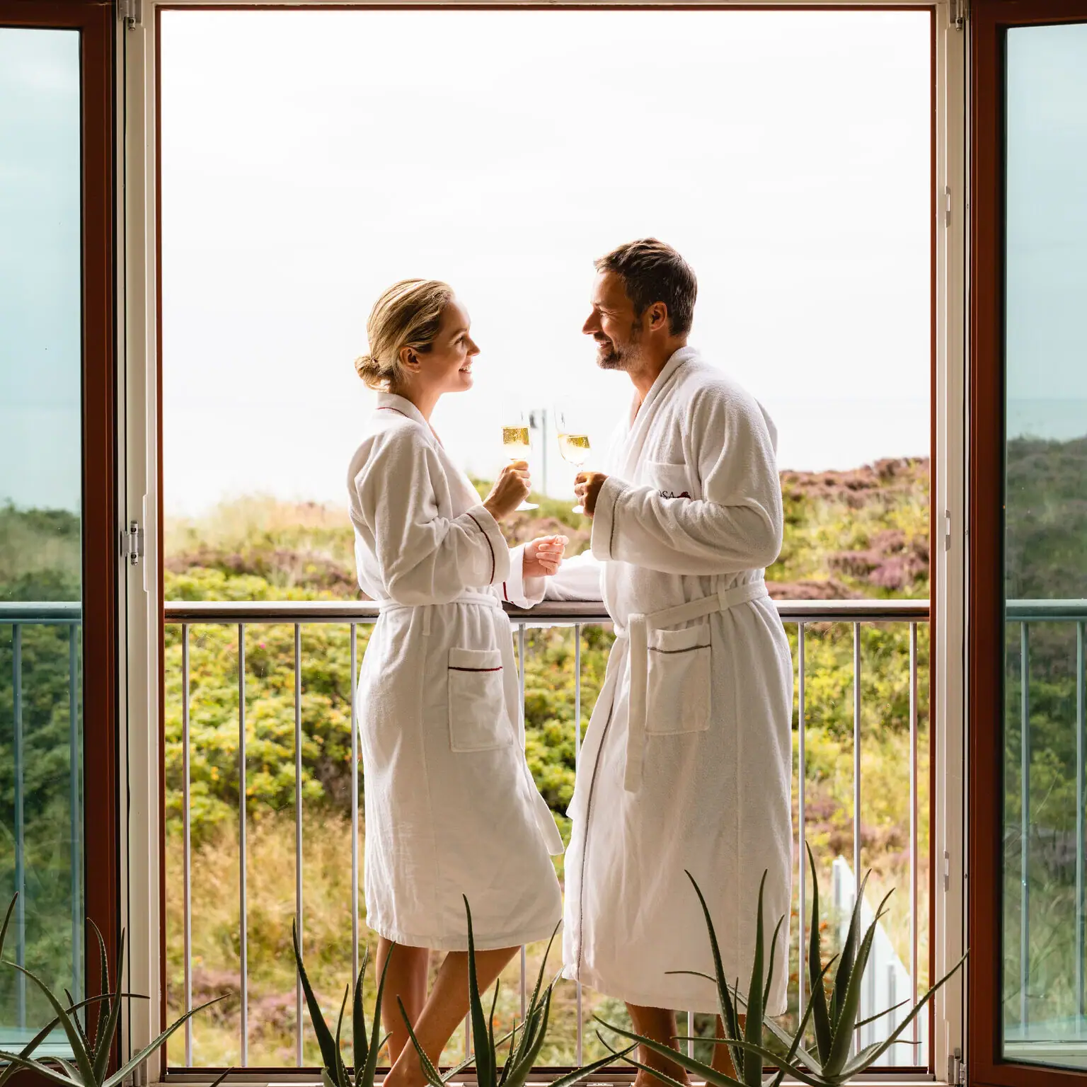 Ein Mann und eine Frau in weißen Bademänteln stehen vor einem Balkon in einer Day SPA Suite im A-ROSA Hotel Sylt.