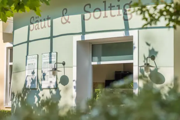 Außenansicht des Bistros „Säut & Soltig“ mit Sonnenschein und grünen Blättern