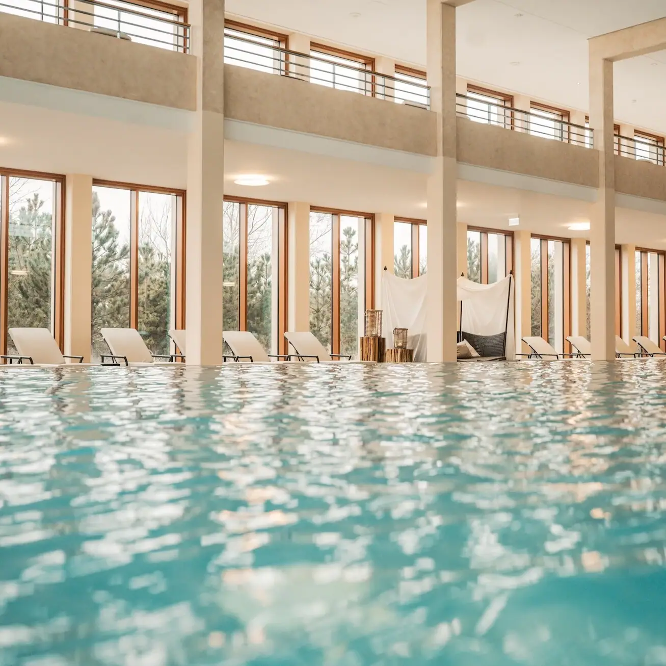 Der Innenpool im A-ROSA Hotel Sylt mit Liegestühlen am Rand und Ausblick auf die Dünenlandschaft durch die Fenster im Hintergrund.
