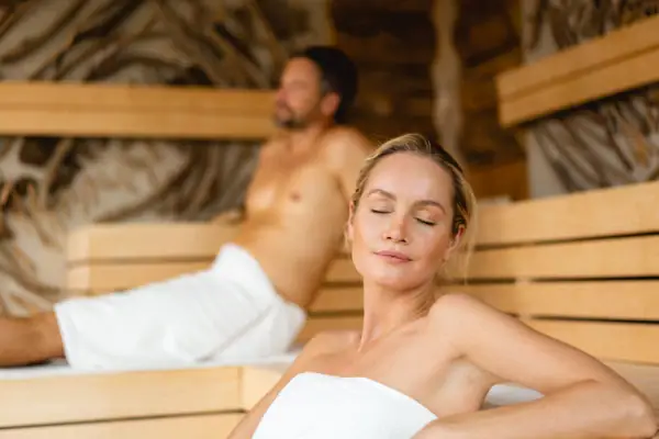 Eine Frau sitzt in einer Sauna, ein Mann im Hintergrund.