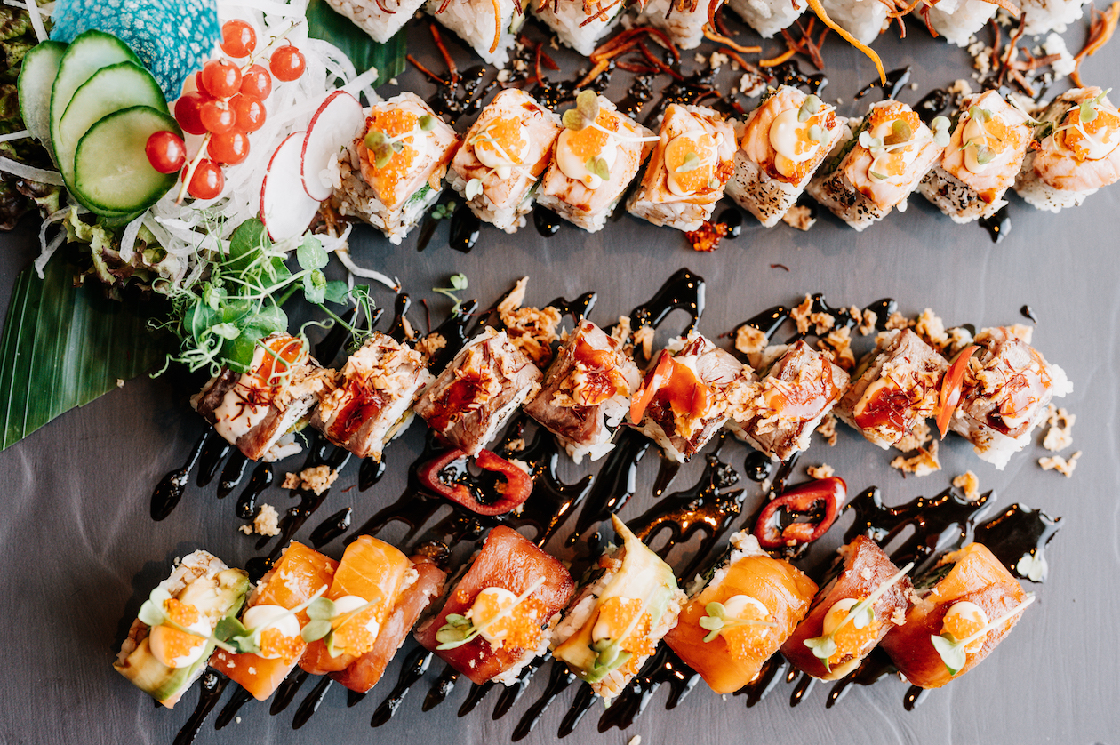 Viele Sushi-Rollen liegen nebeneinander auf einer Schieferplatte und sind aufwendig verziert und angerichtet.