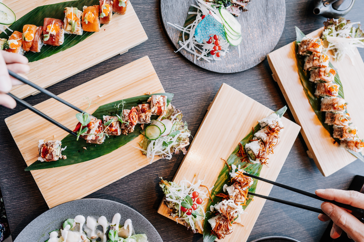 Mehrere Holzbretter mit jeweils einer hübsch angerichteten Sushi-Rolle befinden sich auf einem dunklen Tisch.