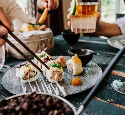 Auf einem dunklen Teller sind einzelne Sushi Häppchen hübsch angerichtete und mit Soße verziert. Eine Hand mit zwei Stäbchen bedient sich daran.