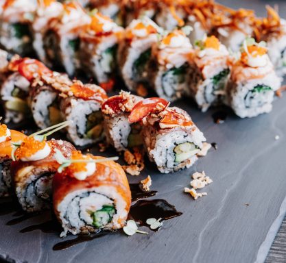 Viele Sushi-Rollen liegen nebeneinander auf einer Schieferplatte und sind aufwendig verziert und angerichtet.